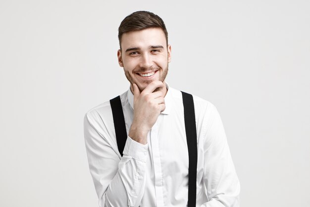 Expresión facial humana positiva, emociones, sentimientos y percepción de la vida. Alegre guapo joven barbudo vestido con elegante camisa blanca con tirantes, riéndose de la broma, frotando su barba