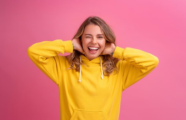 Expresión linda de la cara de la mujer rubia bonita joven que presenta en sudadera con capucha amarilla en fondo rosado brillante aislado emocional divertido