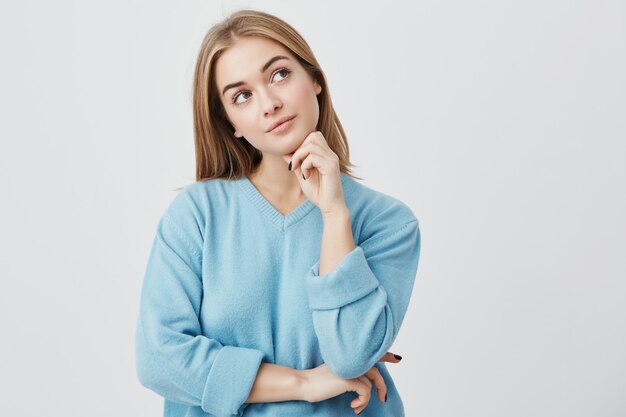 Expresiones faciales y emociones. Chica joven y pensativa con suéter azul con la mano debajo de la cabeza, con una mirada dudosa y sin poder decidir qué ropa ponerse en la fiesta de cumpleaños de un amigo