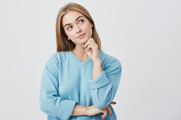 Expresiones faciales y emociones. Chica joven y pensativa con suéter azul con la mano debajo de la cabeza, con una mirada dudosa y sin poder decidir qué ropa ponerse en la fiesta de cumpleaños de un amigo