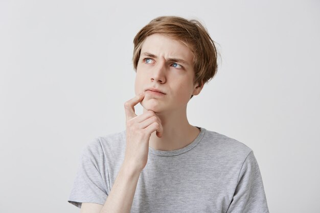 Expresiones faciales, emociones y sentimientos. Joven rubio de ojos azules mirando hacia arriba con expresión pensativa y escéptica, sosteniendo el dedo en la barbilla, tratando de recordar algo importante