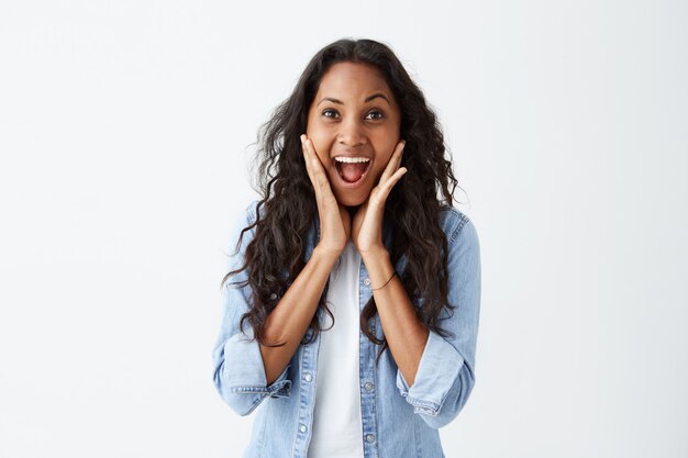 Expresiones faciales humanas, emociones y sentimientos. Mujer morena joven, elegante, de ojos saltones y elegante, con camisa de mezclilla azul que exclama en estado de shock, manteniendo las manos en la cabeza, asombrada con algunas noticias inesperadas