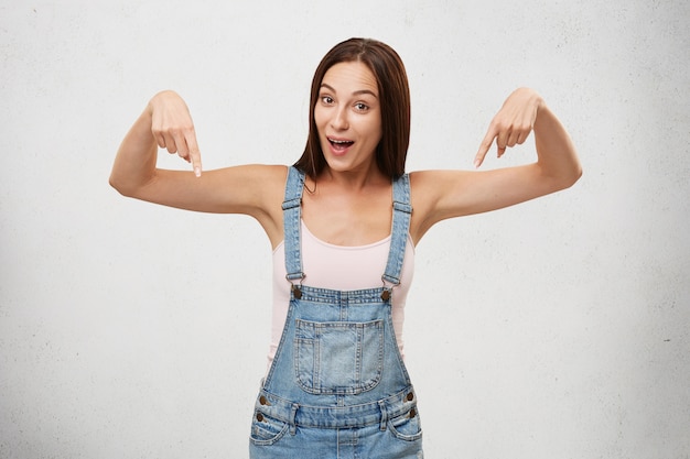 Foto gratuita expresiones faciales humanas positivas. joven mujer emocionada o asombrada de aspecto europeo posando, apuntando con los dedos hacia abajo, exclamando en estado de shock y asombro