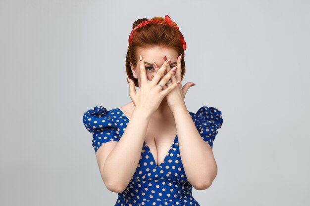 Expresiones faciales humanas, sentimientos y lenguaje corporal. Foto de estudio de asustada jovencita vestida con elegante ropa vintage que cubre la cara con ambas manos, mirando a la cámara a través de los dedos