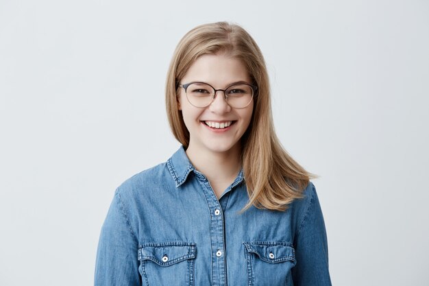 Expresiones faciales positivas y emociones. Atractiva chica estudiante caucásica con cabello rubio y liso en elegantes gafas mirando y sonriendo ampliamente durante una agradable conversación con alguien