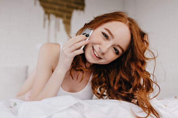 Extasiada chica pelirroja acostada en la cama con teléfono. Modelo femenino europeo romántico que sostiene el teléfono inteligente mientras posa en el dormitorio.