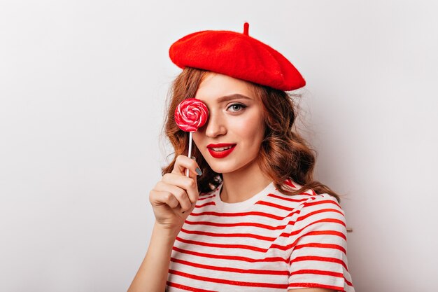 Extasiada chica pelirroja de pie con dulces. Retrato de interior de elegante mujer francesa con piruleta.