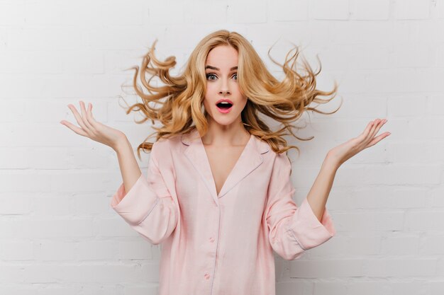 Extasiada mujer de ojos azules con largo cabello rubio posando delante de una pared de ladrillos blancos. Filmación en interiores de una niña sorprendida en un hermoso pijama rosa.