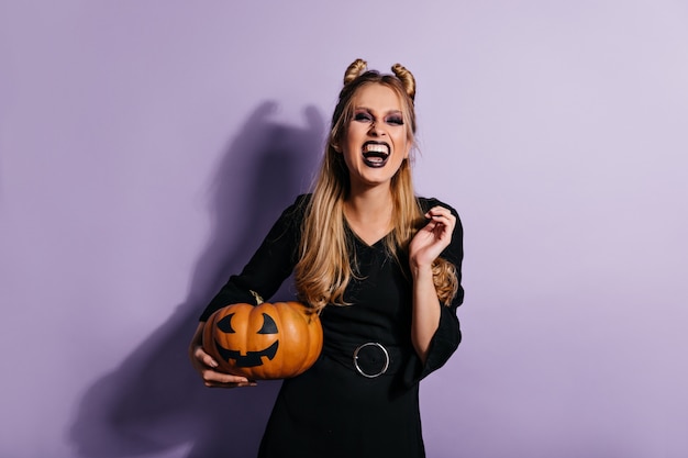 Extasiada mujer de pelo largo con calabaza de halloween y riendo. foto de chica vampiro emocional en vestido negro.