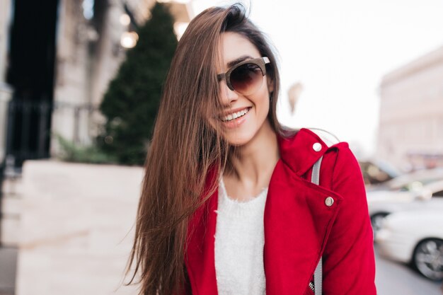 Extasiada mujer de pelo largo en lindas gafas de sol sonriendo en el espacio de la calle