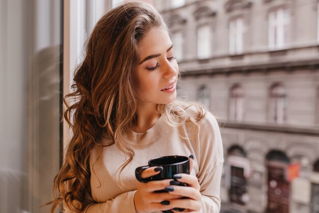 Extasiada mujer rizada sentada con los ojos cerrados cerca de la ventana grande