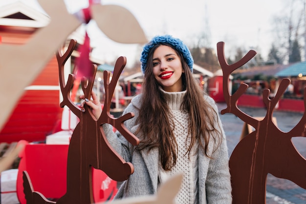 Extasiado modelo femenino de pelo oscuro disfrutando de la Navidad en el parque de atracciones temático. Retrato al aire libre de niña alegre con sombrero azul tejido posando junto a la decoración de vacaciones en invierno.