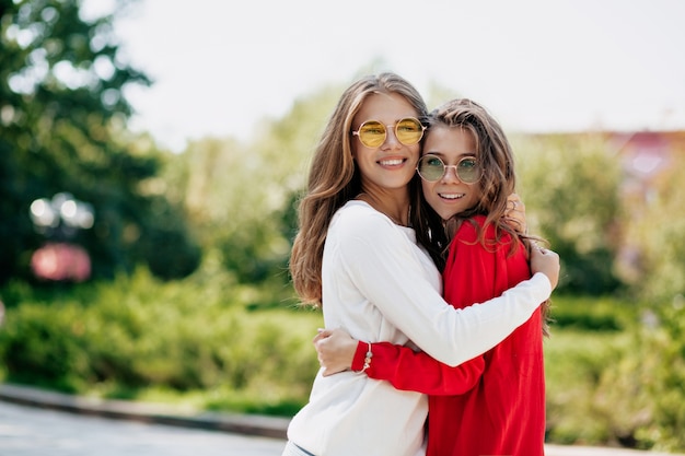 Extasiados mejores amigos felices abrazándose afuera. Mujer joven inspirada vestida con suéteres brillantes y gafas de sol pasar tiempo juntos