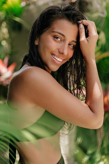 Exterior retrato de atractiva joven mujer bronceada con cabello oscuro y húmedo sonriendo y posando a la cámara sobre fondo de plantas exóticas