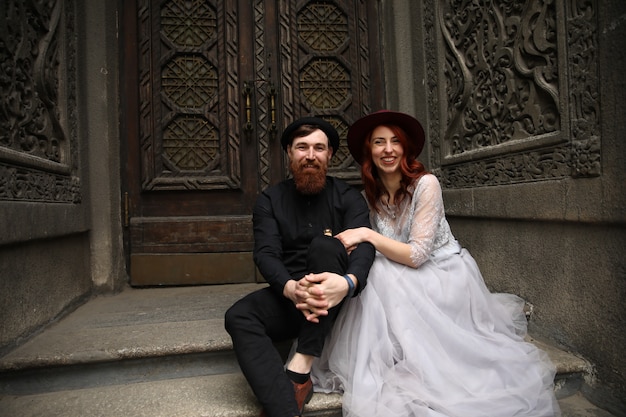 Extraordinaria pareja de novios vestida con sombreros y trajes formales está sentada en las escaleras de piedra y sonriendo