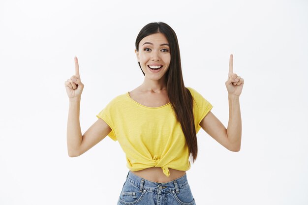 Extrovertida y amistosa divertida chica asiática atractiva con cabello oscuro en camiseta amarilla levantando los dedos índices apuntando hacia arriba con una amplia sonrisa encantada