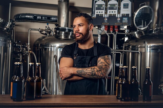 Fabricante masculino hipster tatuado con barba negra que presenta cerveza artesanal en la microcervecería.