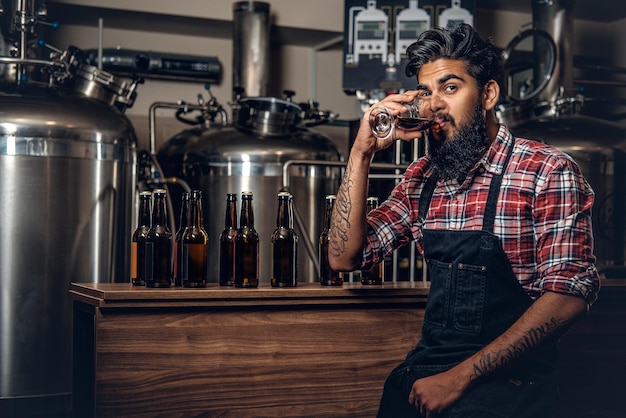 Fabricante masculino hipster tatuado barbudo indio degustando cerveza artesanal en la microcervecería.