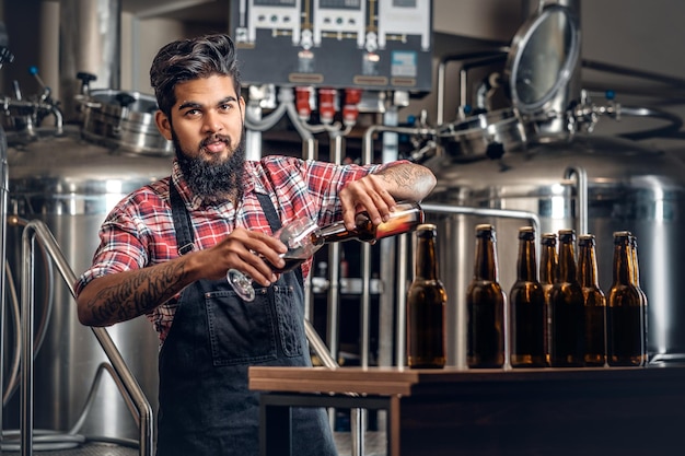Fabricante masculino hipster tatuado barbudo indio degustando y presentando cerveza artesanal en la microcervecería.