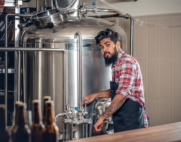 Foto gratuita fabricante masculino hipster tatuado barbudo indio que presenta cerveza artesanal en la microcervecería.