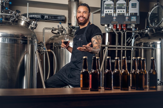 Fabricante masculino hipster tatuado barbudo negro degustando cerveza en la microcervecería.