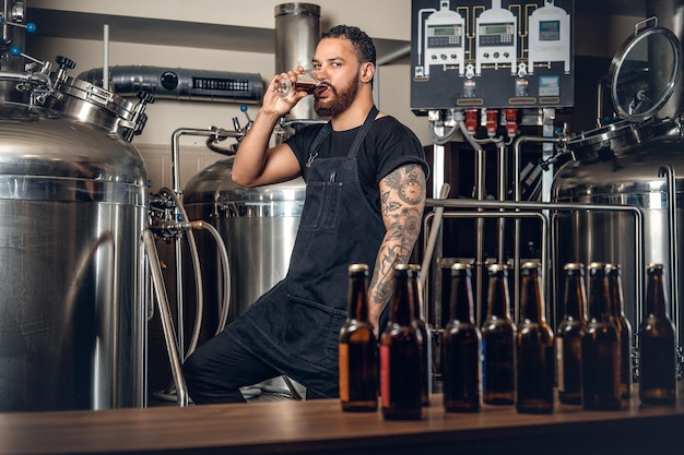 Fabricante masculino hipster tatuado barbudo negro degustando cerveza en la microcervecería.