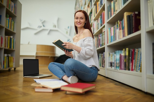 Fácil de aprender. Una joven alegre con el pelo rojo largo y ropa informal con un libro y un bolígrafo sentados en el suelo cerca de una laptop mirando la cámara