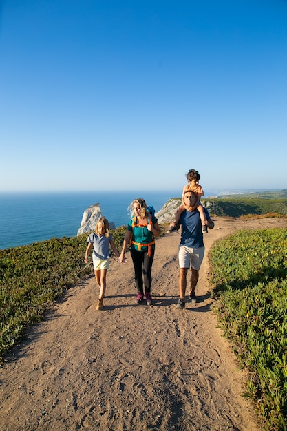 Familia activa pareja e hijos caminando por la orilla del mar, caminando por el sendero. Niño montado en el cuello de los papás. Longitud total. Concepto de naturaleza y recreación