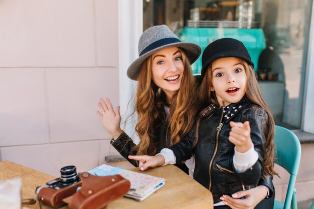 Familia amorosa feliz. Madre e hija sentadas en un café de la ciudad mirando sorprendidas a la cámara y chica mostrando el camino. Sobre la mesa hay un mapa y cámaras. Verdaderas emociones, buen humor ..