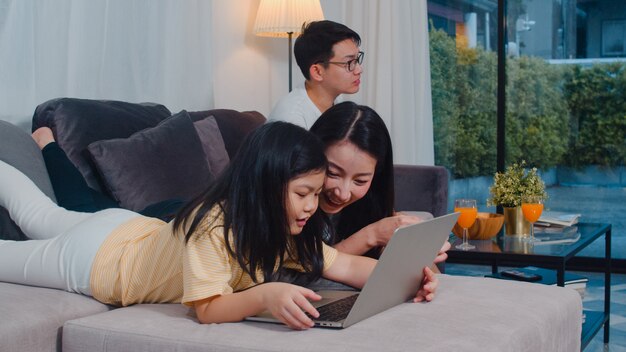 Familia asiática disfruta de su tiempo libre relajarse juntos en casa. Estilo de vida mamá e hija usando una computadora portátil ven películas en internet, papá ven televisión en la sala de estar de una casa moderna.