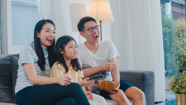 Familia asiática disfruta de su tiempo libre relajarse juntos en casa. Estilo de vida papá, mamá e hija ven la televisión juntos en la sala de estar en la casa moderna por la noche.