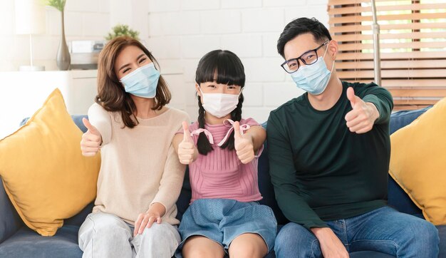 Familia asiática feliz con una máscara para protegerse contra virus y pulgares hacia arriba en la sala de estar de casa