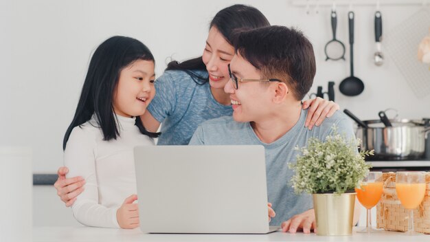 La familia asiática joven disfruta usando la computadora portátil juntos en casa. Estilo de vida joven esposo, esposa e hija feliz abrazo y jugar después de desayunar en la cocina moderna en casa por la mañana.
