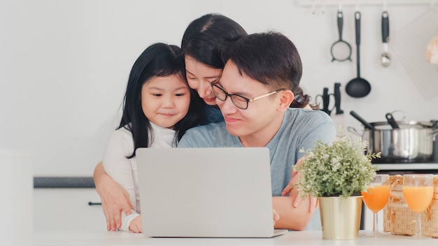 La familia asiática joven disfruta usando la computadora portátil juntos en casa. Estilo de vida joven esposo, esposa e hija feliz abrazo y jugar después de desayunar en la cocina moderna en casa por la mañana.