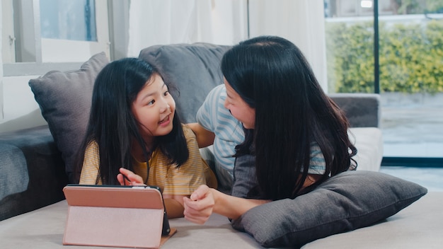 Familia asiática joven e hija felices usando la tableta en casa. Madre japonesa relajarse con niña viendo la película acostado en el sofá en la sala de estar en casa. Mamá divertida y niño encantador se divierten.