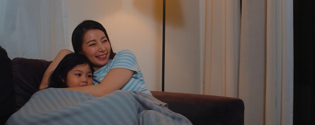 Familia asiática joven e hija viendo la televisión en casa en la noche. madre coreana con niña feliz usando el tiempo en familia relajarse tumbado en el sofá en la sala de estar. mamá divertida y niño encantador se divierten.