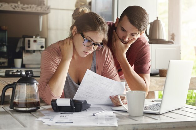 Familia caucásica joven que tiene problemas de deuda, no puede pagar su préstamo. Mujer con gafas y hombre morena estudiando banco de formularios en papel mientras gestionan el presupuesto doméstico juntos en el interior de la cocina