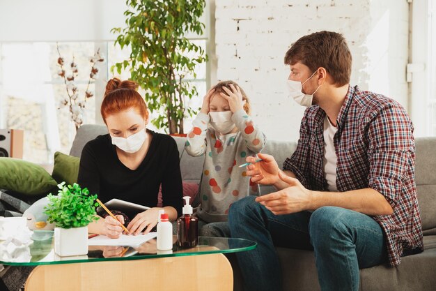 Familia caucásica en mascarillas y guantes aislados en casa con síntomas respiratorios por coronavirus como fiebre, dolor de cabeza, tos en estado leve. Cuidado de la salud, medicina, cuarentena, concepto de tratamiento.