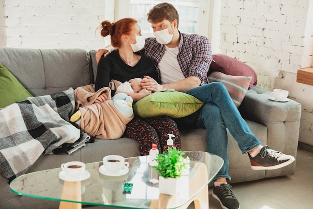 Familia caucásica en mascarillas y guantes aislados en casa con síntomas respiratorios de coronavirus como fiebre, dolor de cabeza, tos en estado leve.