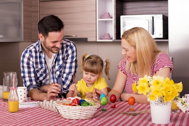 Familia caucásica sentada detrás de la encimera de la cocina con su hija y pintando huevos de Pascua