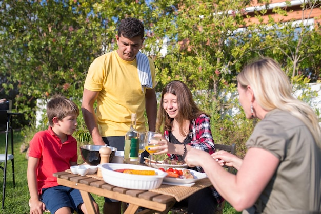 Familia feliz comiendo barbacoa en el patio trasero. Padres e hijos adultos de mediana edad sentados alrededor de la mesa, comiendo alimentos recién cocinados, bebiendo bebidas. Barbacoa, cocina, comida, concepto familiar.