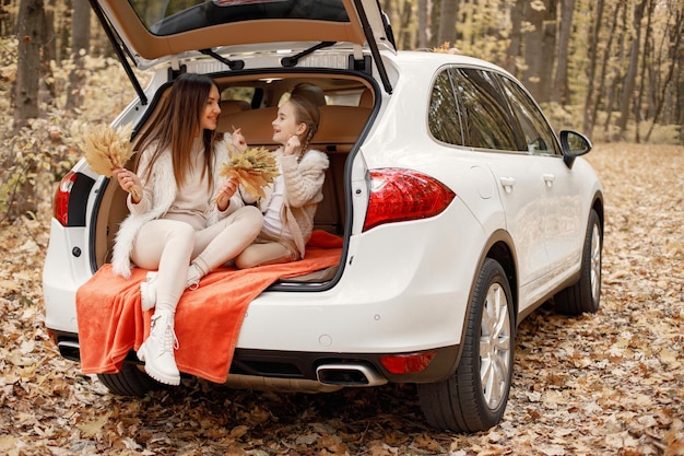 Familia feliz descansando después de pasar el día al aire libre en el parque de otoño. La madre y su niña sentada dentro del baúl del auto blanco. Madre e hija vestidas de blanco.