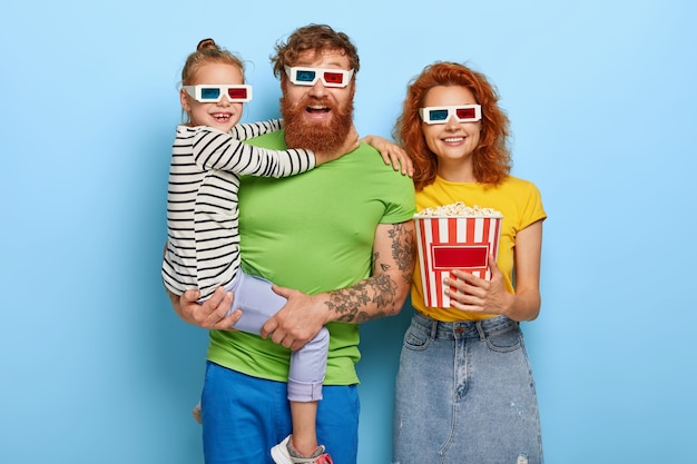 La familia feliz disfruta de películas o dibujos animados en el cine, usa gafas 3D, se divierte con efectos visuales y de sonido geniales, come un delicioso refrigerio. La pequeña niña en manos de los padres, lo abraza. Gente, ocio, fin de semana