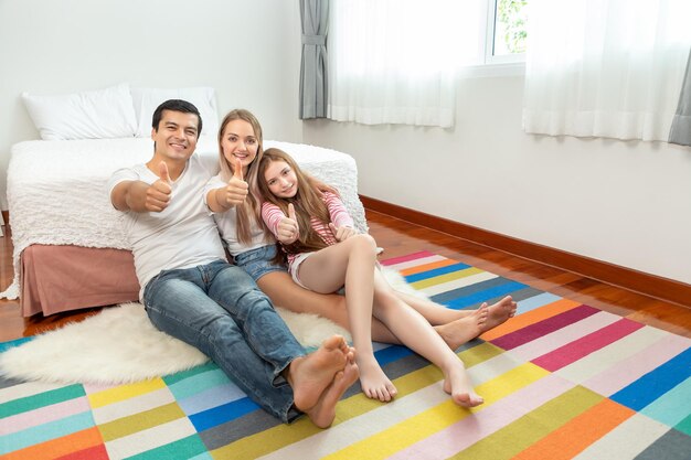 La familia feliz, incluido el padre, la madre y la hija, están sentados frente a la cama con una sonrisa y el pulgar hacia arriba mirando a la cámara