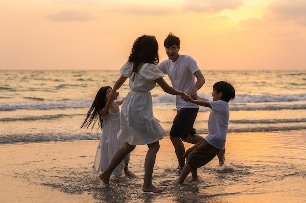 La familia feliz joven asiática disfruta de vacaciones en la playa en la noche. Papá, mamá y niño se relajan jugando juntos cerca del mar cuando la puesta de sol silueta Concepto de estilo de vida viajes vacaciones vacaciones verano.