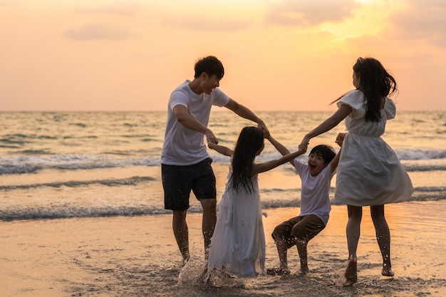 La familia feliz joven asiática disfruta de vacaciones en la playa en la noche. Papá, mamá y niño se relajan jugando juntos cerca del mar cuando la puesta de sol silueta Concepto de estilo de vida viajes vacaciones vacaciones verano.
