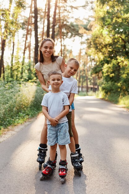 Familia feliz, mujer de cabello oscuro con ropa casual de pie con sus hijos al aire libre, madre con niños patinando en el parque en la carretera asfaltada, divirtiéndose juntos.