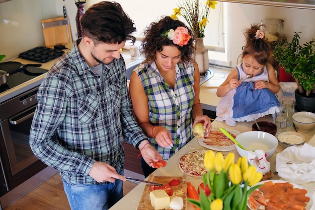 Familia feliz de mujer morena, hombre guapo con una camisa de lana y una linda niña cocinando comida en una cocina casera.