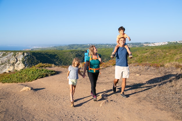 Familia feliz pareja y niños senderismo en el campo, caminando por el sendero. Muchacho emocionado montando en el cuello de los papás. Longitud total. Concepto de naturaleza y recreación