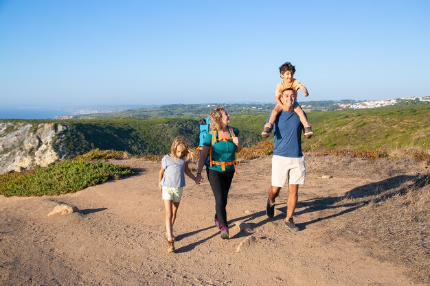 Familia feliz pareja y niños senderismo en el campo, caminando por el sendero. Muchacho emocionado montando en el cuello de los papás. Longitud total. Concepto de naturaleza y recreación